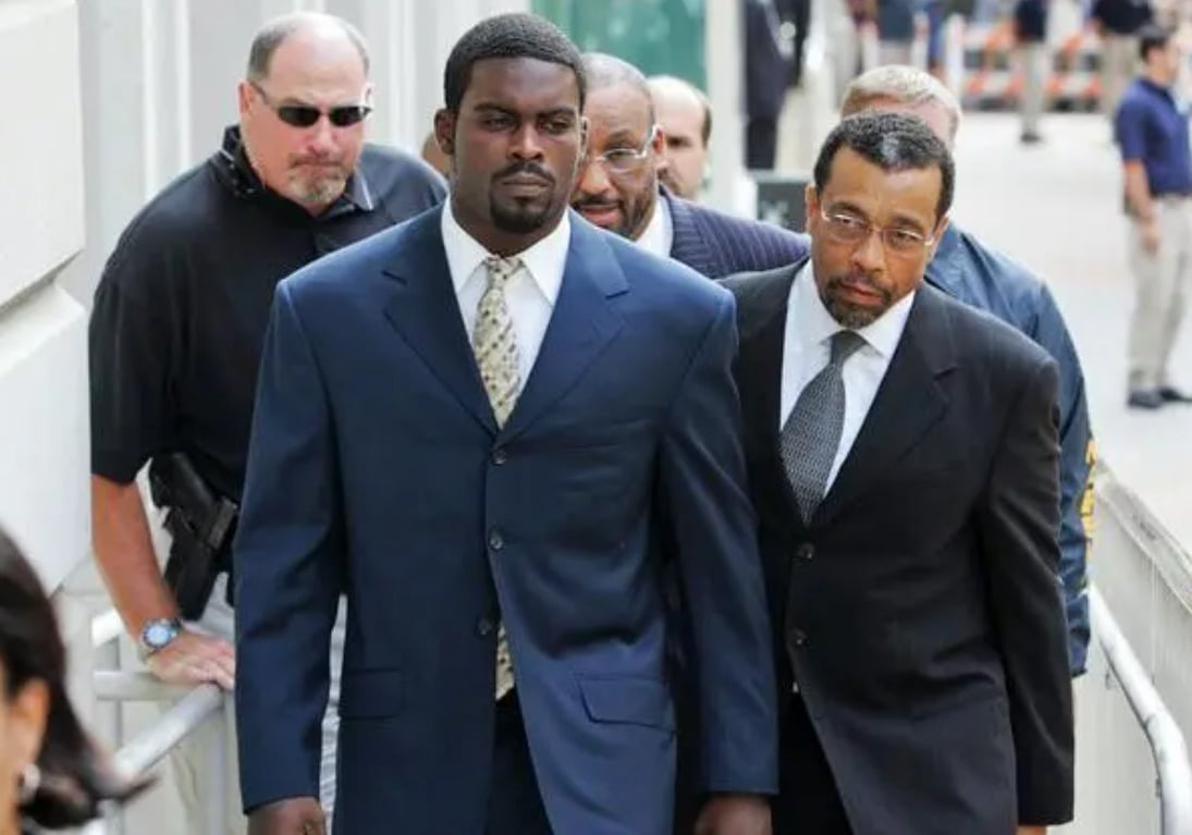 A man in a dark suit walks outside alongside a lawyer and several others, with security personnel nearby and people in the background.