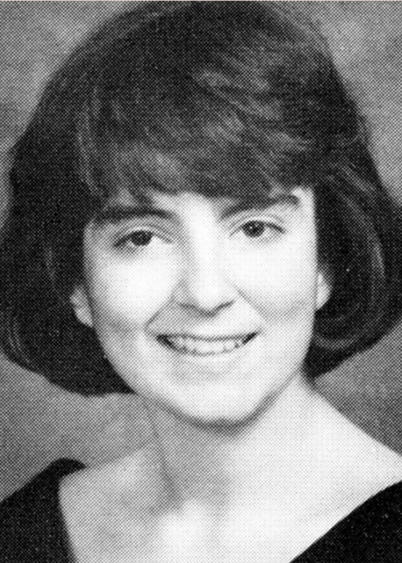 Black and white portrait of a young woman with short, dark hair, wearing a dark off-the-shoulder top, smiling gently at the camera against a plain studio background.