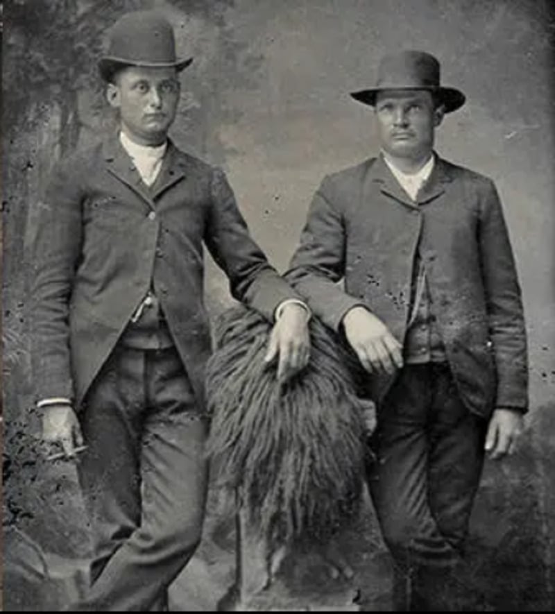 Two men in old-fashioned suits and bowler hats pose for a black-and-white vintage photograph, each resting an arm on a small table draped with a fringed cloth. The background features a painted outdoor scene.