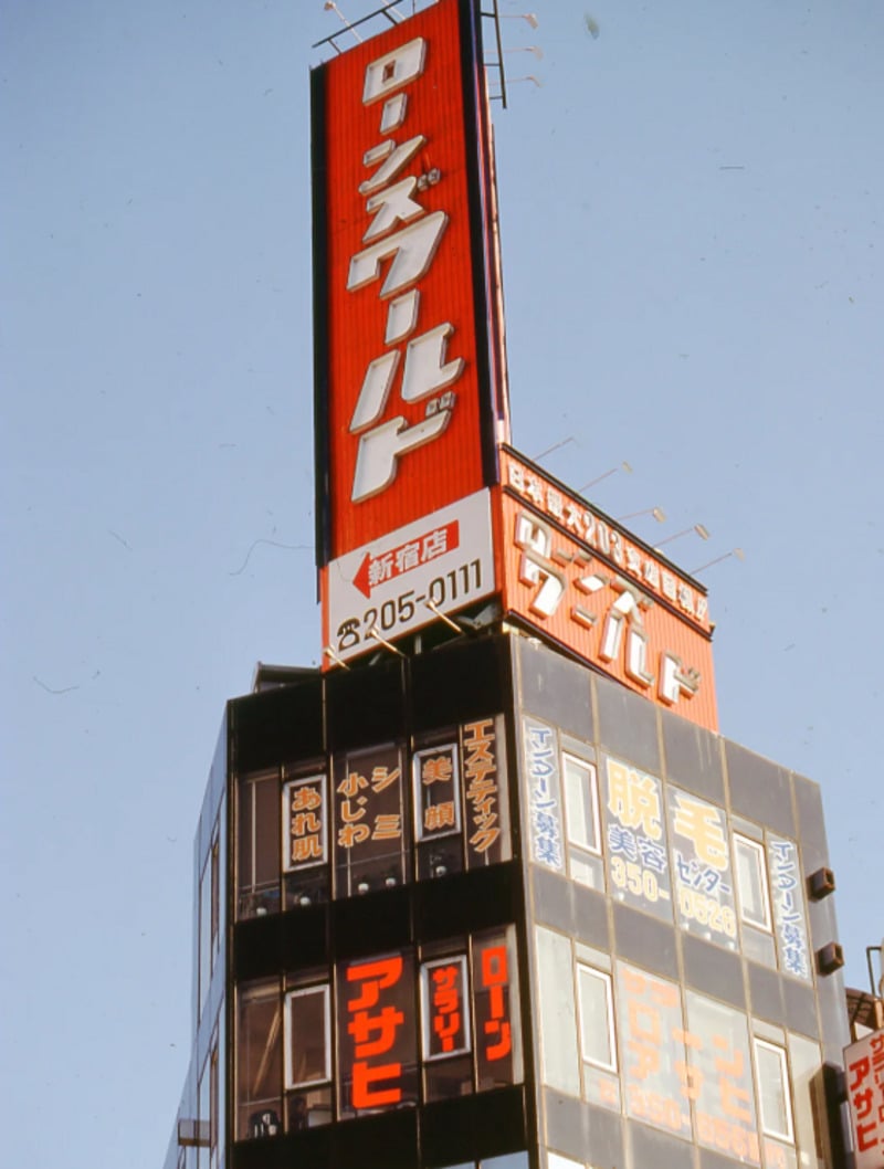 A tall building in Japan with large red and white vertical signs displaying Japanese text, set against a clear blue sky. The building has glass windows and multiple illuminated advertisements.