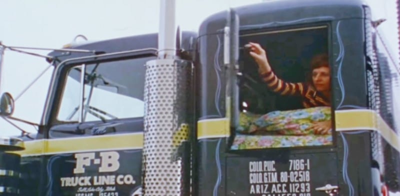 A person in a striped shirt leans out of the window of a dark blue F-B Truck Line Co. semi-truck, reaching upward with one hand. Colorful bedding is visible inside the truck's cab.