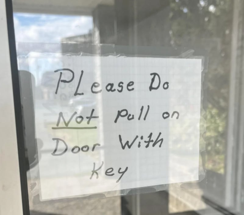 A handwritten sign taped to a glass door reads, "Please Do Not pull on Door With Key," with "Not" underlined for emphasis. Sunlight and outdoor reflections are visible on the glass.