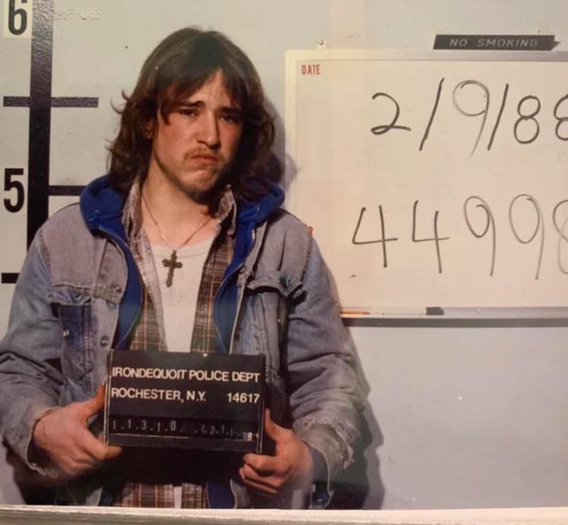 A young man with long hair, wearing a denim jacket and cross necklace, stands for a mugshot. He holds a sign reading "Irondequoit Police Dept, Rochester, N.Y." with a date and numbers written on a board behind him.