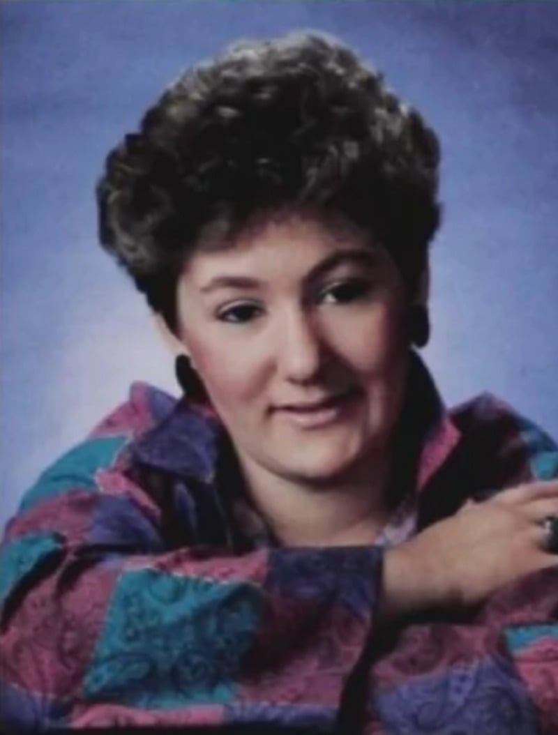 A person with short, curly brown hair is wearing a colorful, patterned shirt and smiling gently, resting one arm on a surface against a purple backdrop.