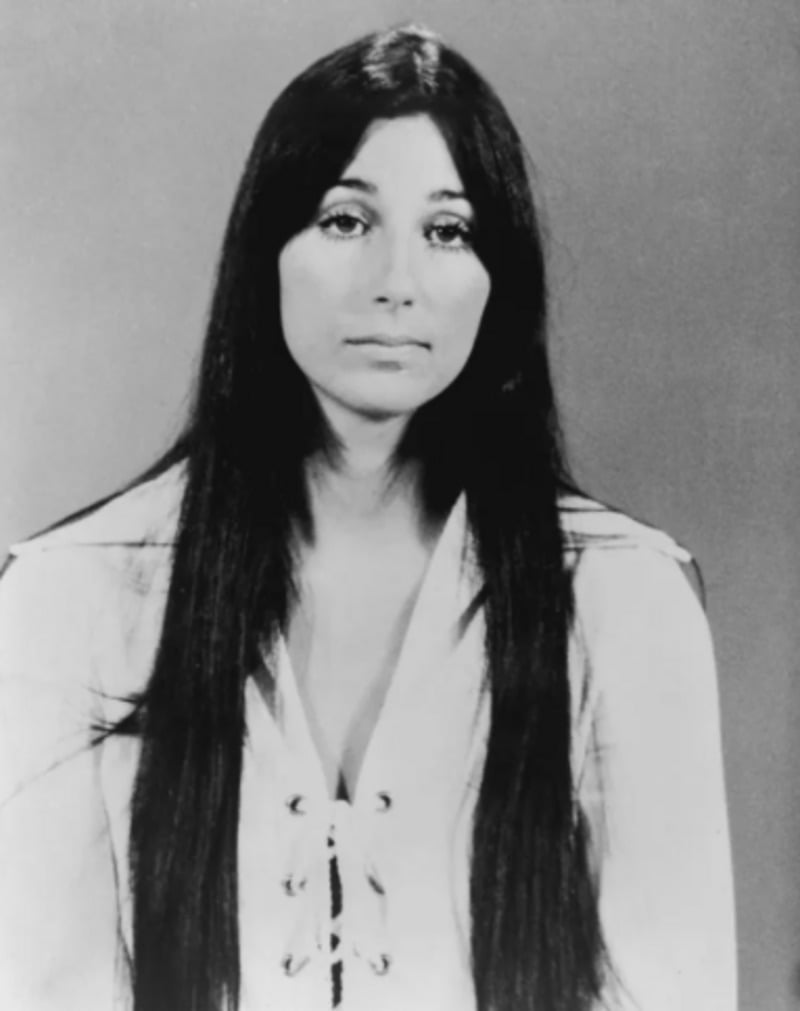 Black-and-white portrait of a woman with long straight dark hair, wearing a light-colored lace-up top, looking directly at the camera with a neutral expression against a plain background.