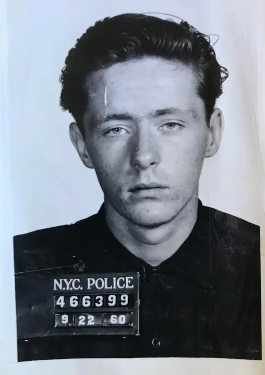 Black-and-white mugshot of a young man with short, dark hair, wearing a dark shirt. A placard reading "N.Y.C. Police 466399 9 22 60" is displayed on his chest.