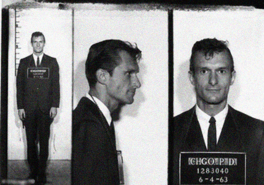 Black-and-white mugshot featuring a young man in a suit, shown full body, profile, and front view. He holds a sign displaying "CHGO. P.D. 1283040, 6-4-63" and stands against a height chart.