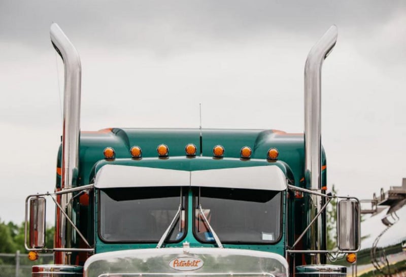 A close-up front view of a green Peterbilt semi-truck with tall chrome exhaust stacks and orange lights on top, parked outdoors under a cloudy sky.