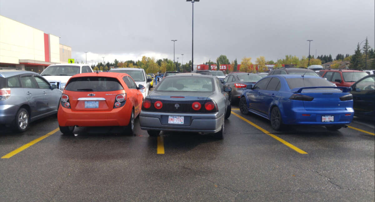 Three cars are parked in a parking lot; the gray car in the middle is parked over the line, taking up two spaces, between an orange car on the left and a blue car on the right.