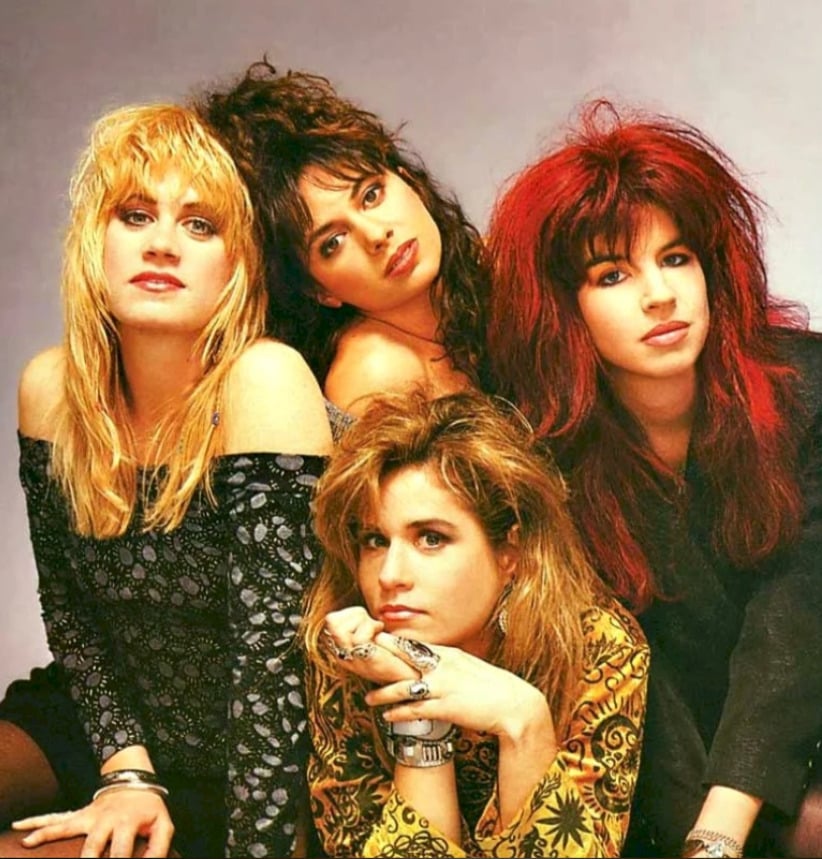 Four women with teased, colorful 1980s hairstyles and vibrant clothing pose together against a neutral background, giving confident and expressive looks to the camera.