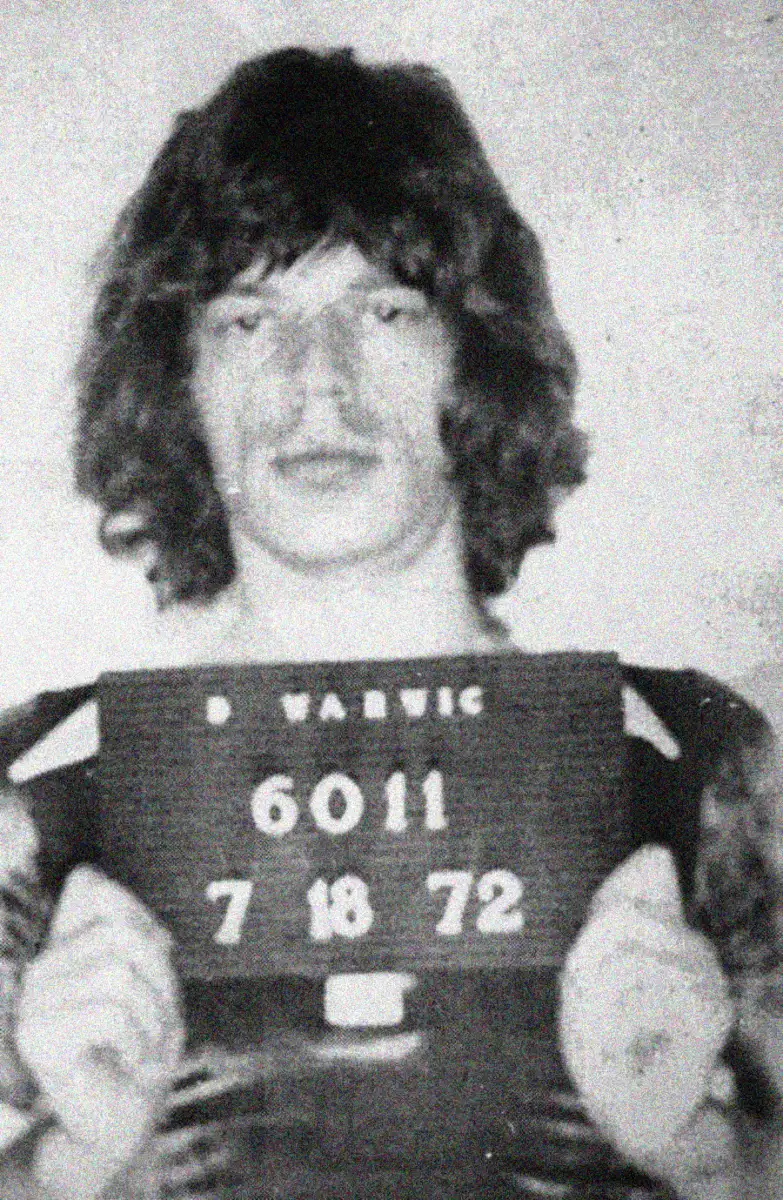 Black and white photo of a person holding a sign with the text "B WARWIC 6011 7 18 72", resembling a mugshot. The person has medium-length wavy hair and is looking at the camera.