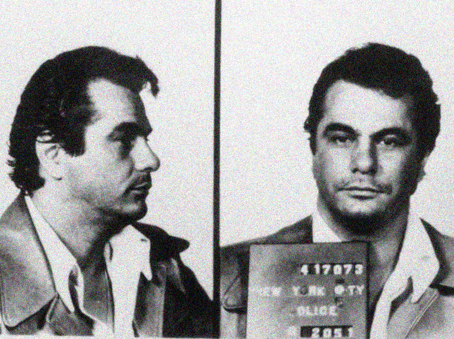 Black and white mugshot showing a man with dark hair in a collared shirt and jacket, photographed from the side and front, holding a New York City police placard.