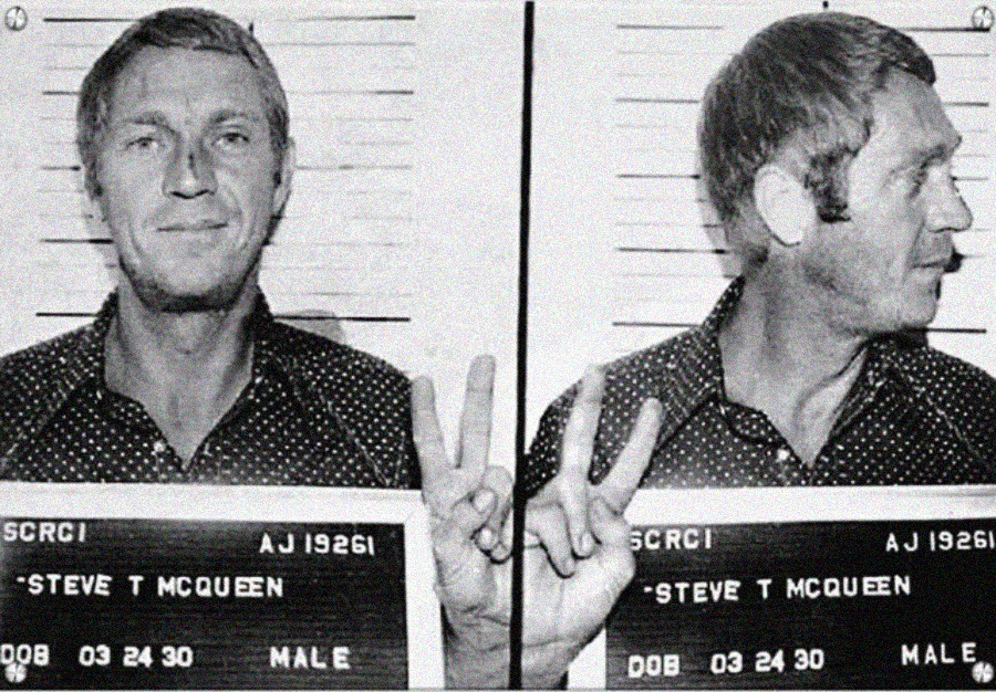 Black and white mugshot of a man holding a sign that reads “Steve T McQueen.” He is smiling and making a peace sign with his fingers in both front and side profile photos against a height chart background.