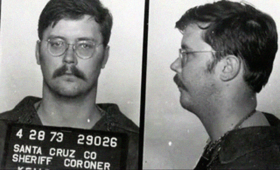 Black and white mugshot of a man with glasses and a mustache, shown from the front and side. He is holding a sign that reads "Santa Cruz Co Sheriff Coroner" with identification numbers.