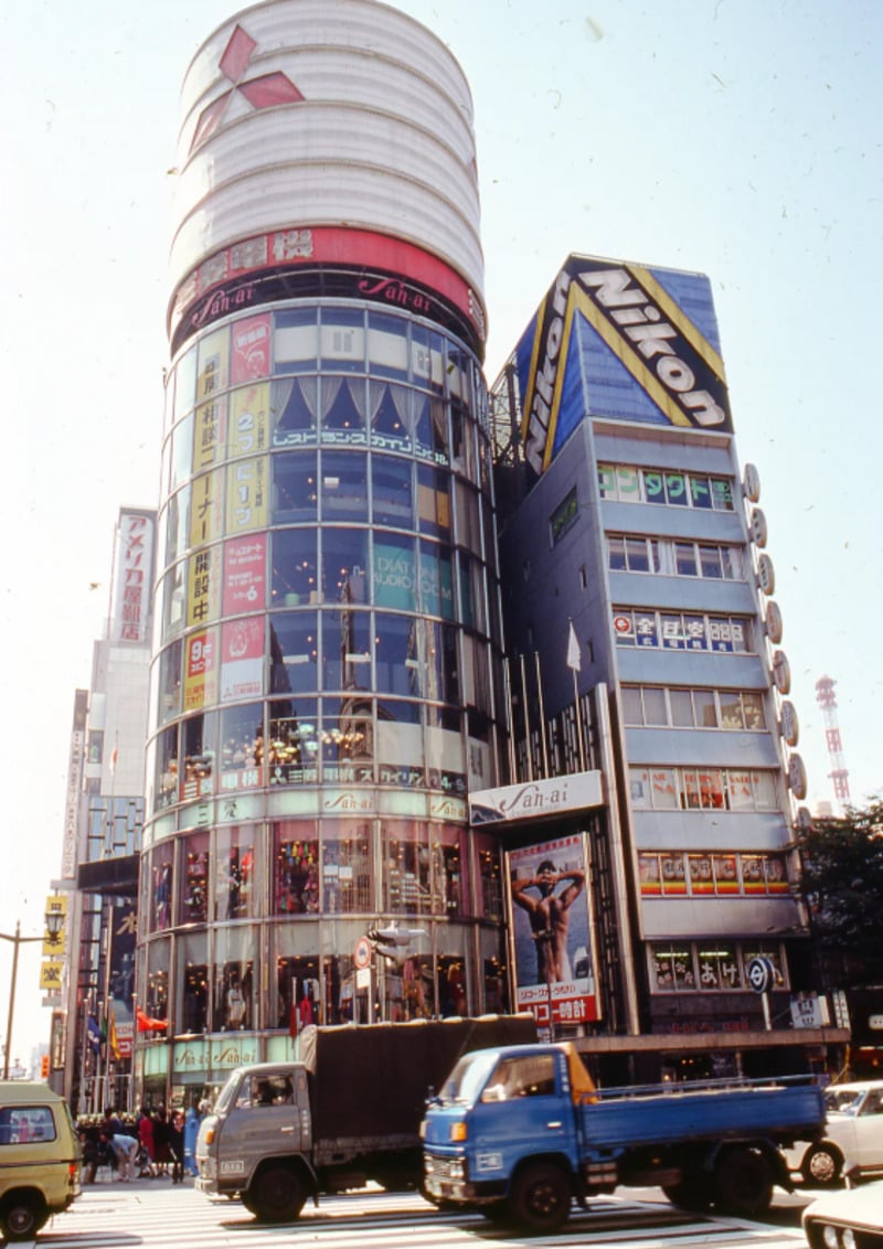 A busy city street with trucks passing in front of tall, glass-fronted buildings. One building features a large "Nikon" sign and colorful advertisements in Japanese, while the other displays a circular top and the "Saha" logo.