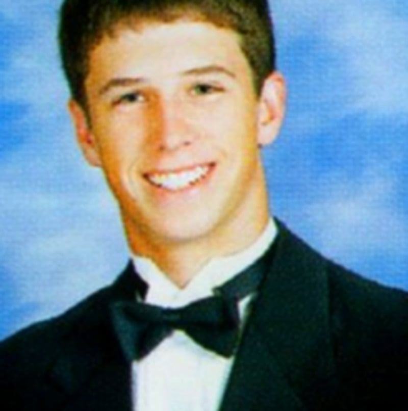 A young man with short brown hair smiles in a formal studio portrait. He is wearing a black tuxedo jacket, white shirt, and black bow tie, with a blue and white cloudy background.