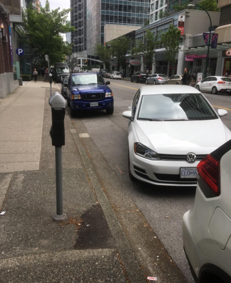 A city street with parked cars along the curb, including a white Volkswagen and a blue truck. Buildings and trees line the sidewalk, and a parking meter is visible in the foreground.