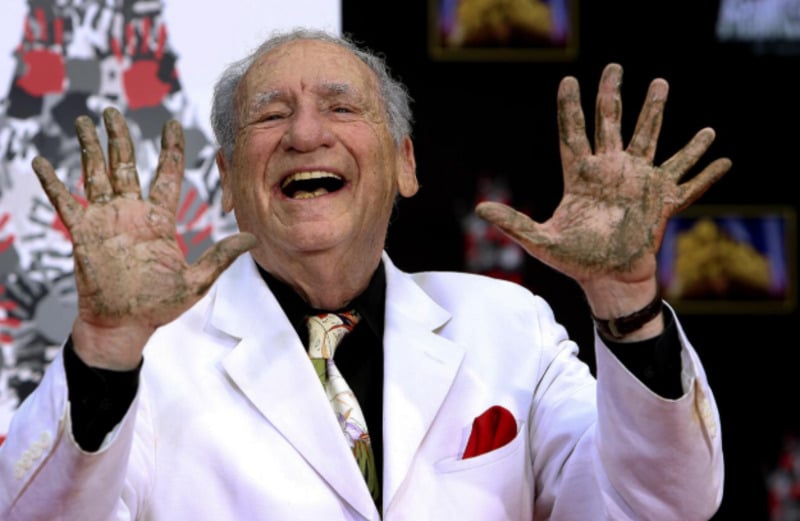 An older man in a white suit smiles broadly, holding up his hands covered in wet cement, likely at a handprint ceremony.