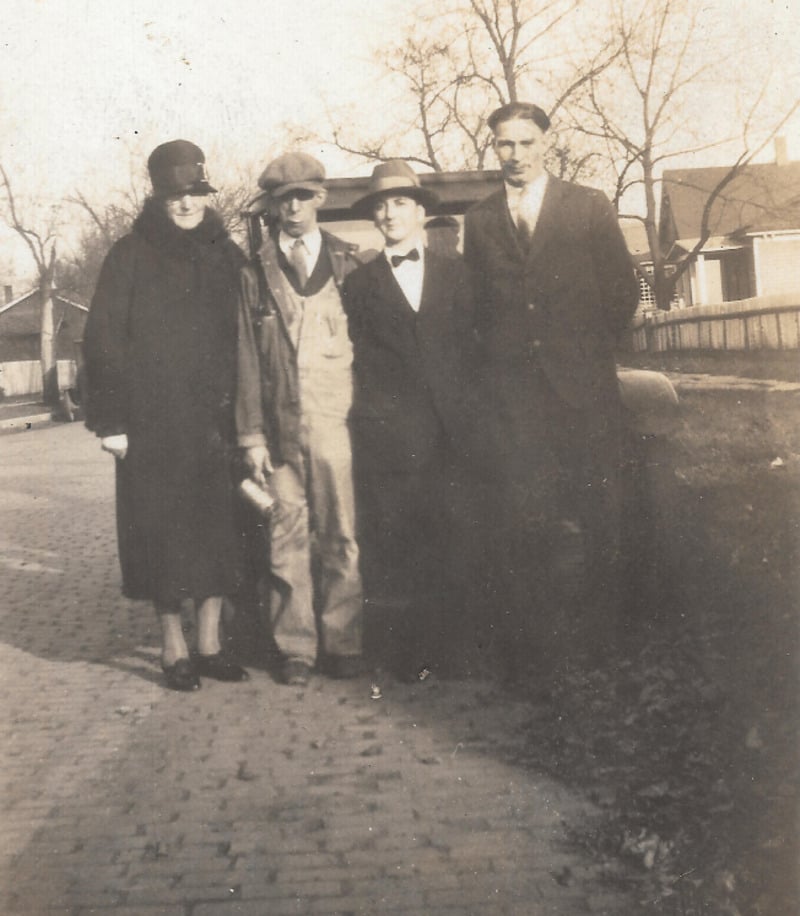 Four people stand on a cobblestone street in vintage clothing. Two men wear suits and hats, one man wears overalls, and a woman wears a long coat and cloche hat. Leafless trees and houses appear in the background.