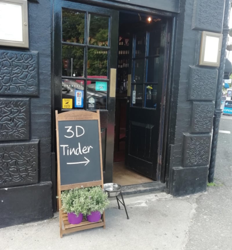 A blackboard sign outside a dark-painted building reads "3D Tinder →" with two potted plants nearby. The open door reveals part of the interior, and a water bowl sits on the ground beside the entrance.