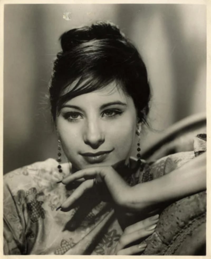 A young woman with dark hair styled up rests her chin on her hand and gazes thoughtfully. She wears dangling earrings and a patterned top, with a soft-focus background.