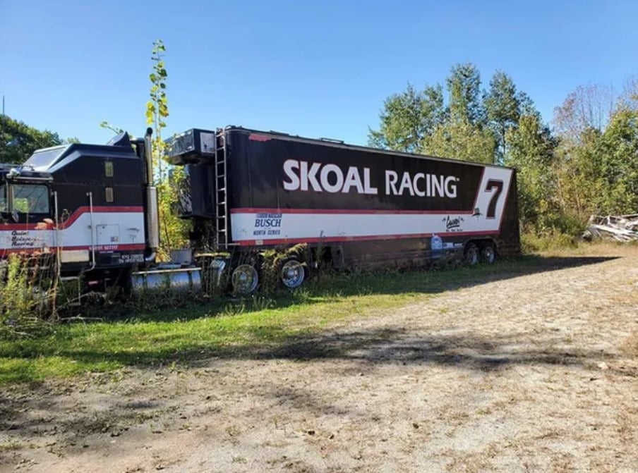 A black and white semi-truck trailer with "SKOAL RACING #7" branding sits unused on grass near a dirt road, surrounded by greenery and tall trees under a clear blue sky.