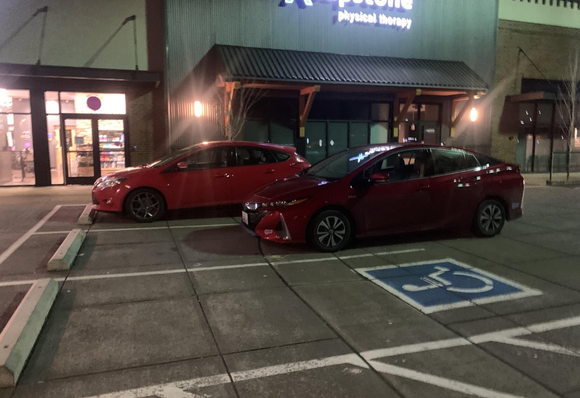 Two red cars are parked outside a building at night. One car is parked in a handicap spot without a visible placard, while the other is in a regular spot. The building has a sign that reads "physical therapy.
