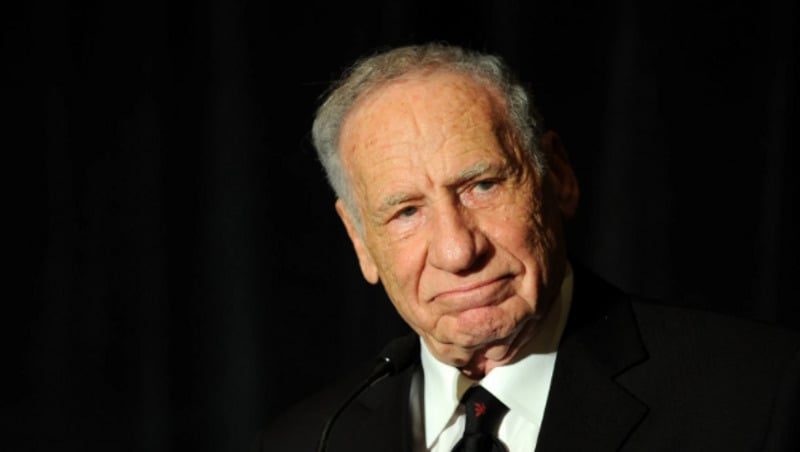 An older man in a dark suit and tie stands in front of a dark background, looking slightly to the side with a thoughtful expression. He has short gray hair and is illuminated by a soft light.