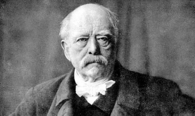 An older man with a bald head, bushy eyebrows, and a large mustache wearing a dark suit and a white bow tie, looking seriously at the camera. The image is black and white.