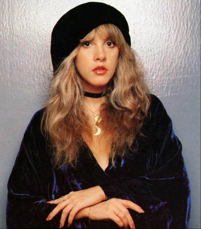 A woman with long, wavy blonde hair wears a black beret, a dark velvet top, and a crescent moon necklace, standing against a plain, light-colored background.