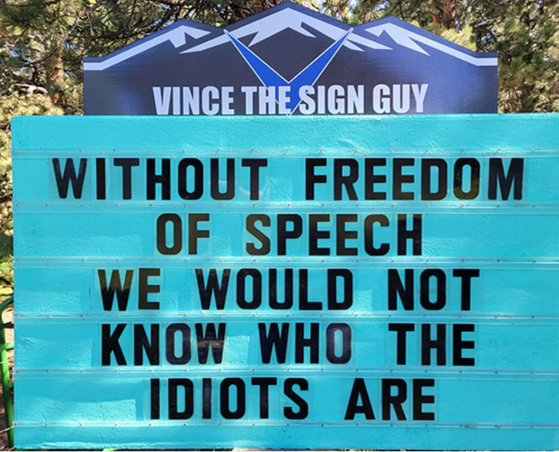 A teal sign reads: “WITHOUT FREEDOM OF SPEECH WE WOULD NOT KNOW WHO THE IDIOTS ARE.” The sign is under a banner that says “VINCE THE SIGN GUY” with a mountain graphic in the background.