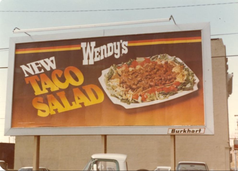 A vintage billboard advertises Wendy's new Taco Salad, featuring a large photo of the salad with beef, lettuce, and tomatoes on a white plate. Bold yellow and orange letters promote the item against a dark background.