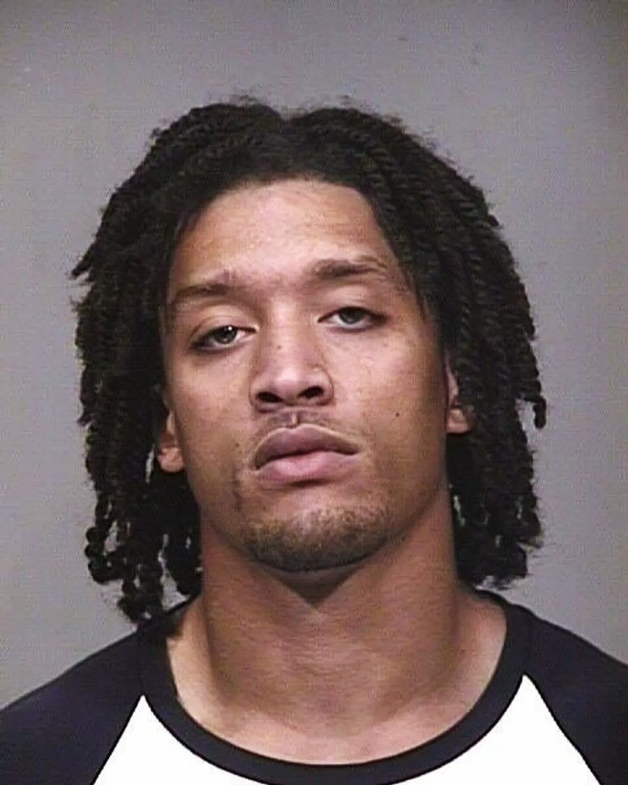A man with medium-length twisted hair, slight facial hair, and neutral expression faces the camera against a plain gray background, wearing a black and white shirt.