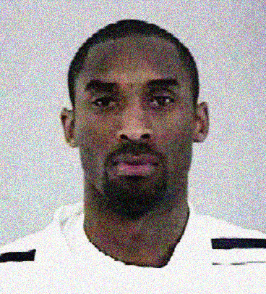 A man with short hair and a goatee is facing forward, wearing a white shirt with black stripes on the shoulders, against a plain light-colored background.