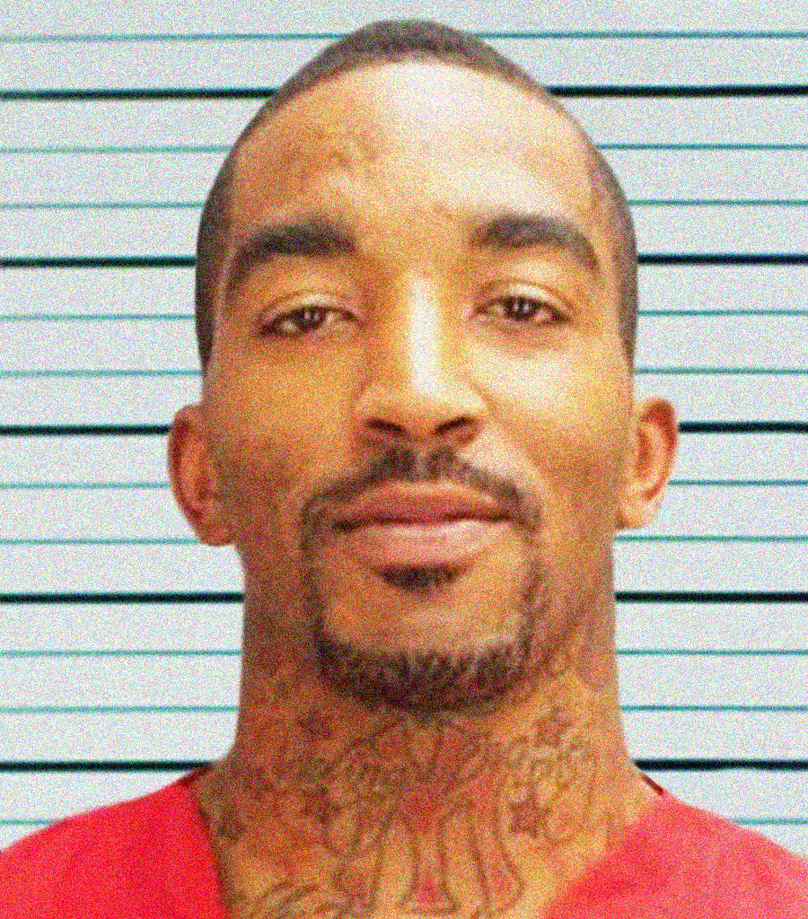 A man with short hair, goatee, and neck tattoos wears a red shirt, posing in front of a height chart with horizontal black lines.