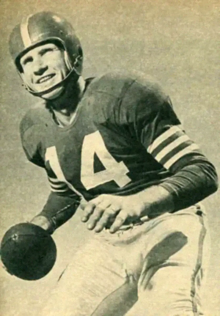 Black-and-white photo of a smiling American football player wearing a helmet and a jersey with the number 14, poised to throw a football. The background is plain, emphasizing the player.