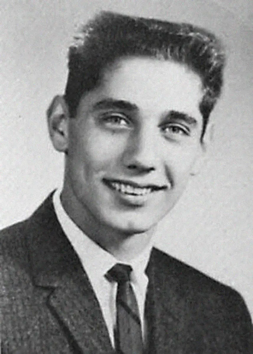 A young man with short dark hair smiles while wearing a suit and tie in a black-and-white portrait photograph.