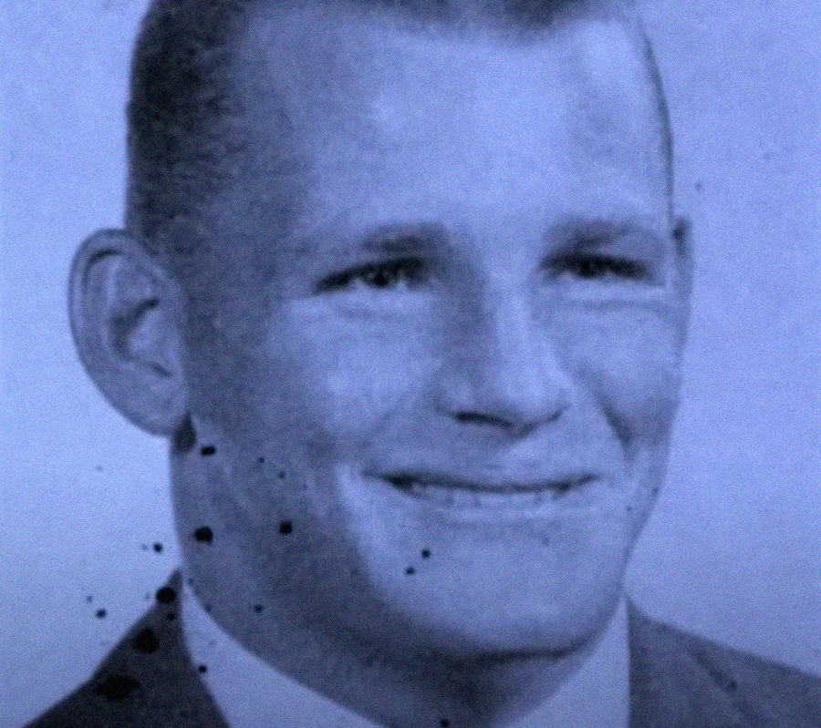 A black and white portrait photo of a man in a suit, smiling, with short hair. The image has several dark specks and marks, giving it an aged or damaged appearance.
