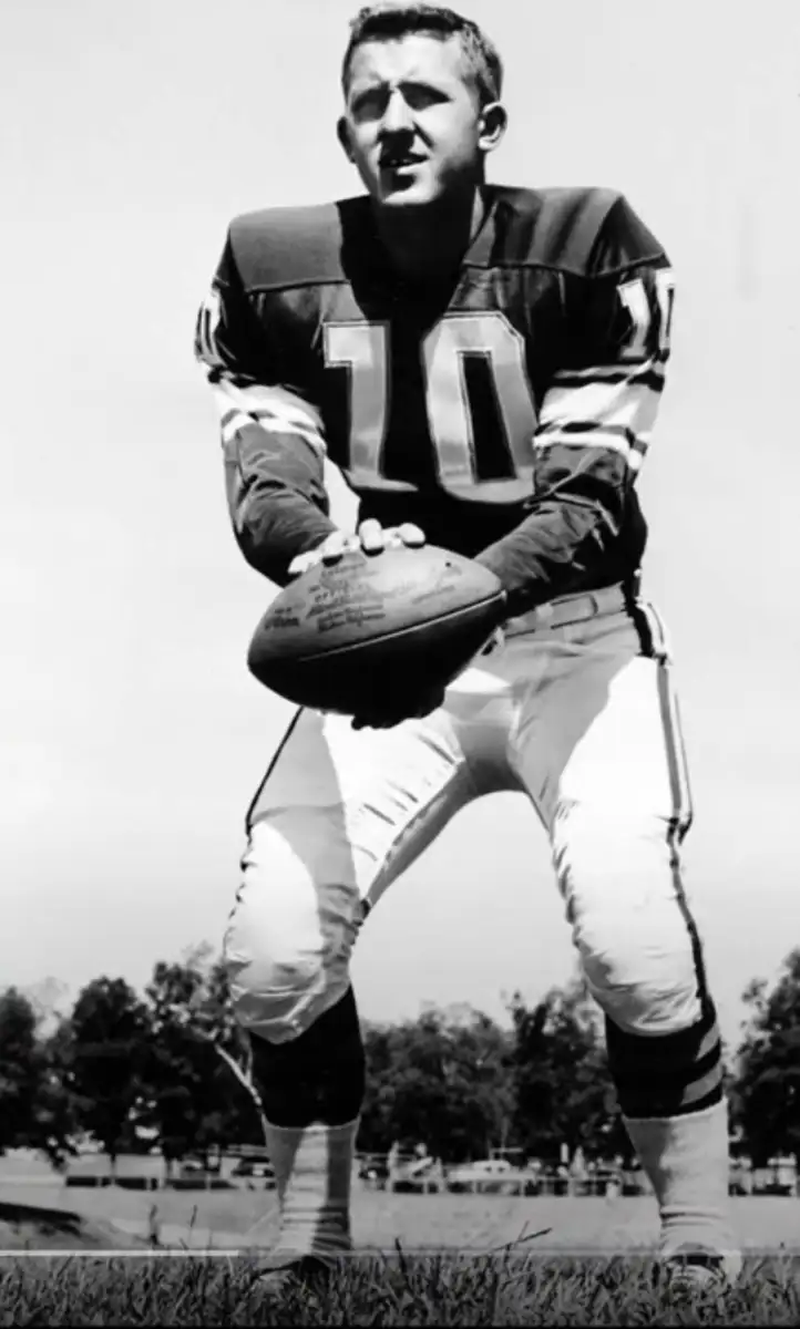 A black-and-white photo of a football player in a number 10 jersey holding a football, crouched in a ready position outdoors with trees visible in the background.