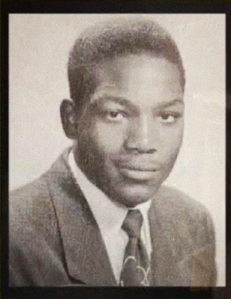Black and white portrait of a young man wearing a suit and tie, looking at the camera with a neutral expression. The background is plain and light-colored.