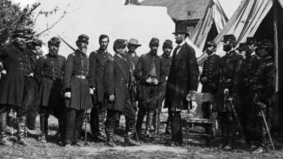 A group of men in military uniforms stand outdoors with a tall man in a suit and stovepipe hat, likely from the 19th century, near tents and a white backdrop. Antietam