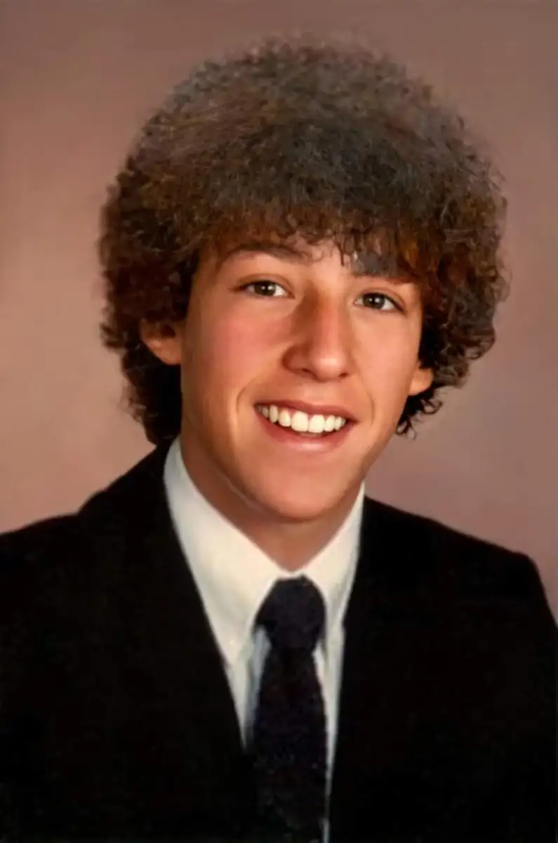 A young man with curly brown hair wearing a black suit, white shirt, and dark tie smiles at the camera against a plain, softly blurred background.