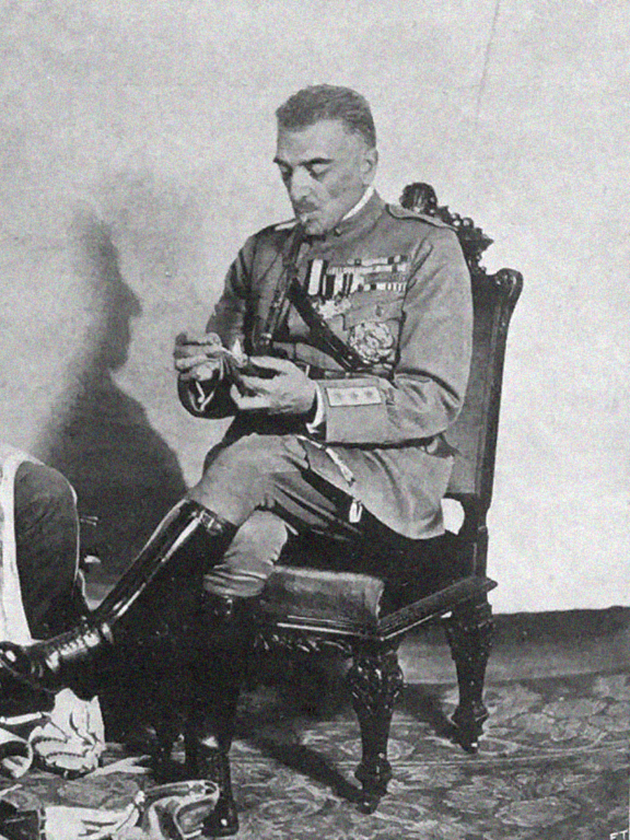 A man in a decorated military uniform sits on an ornate chair, looking down and handling an object in his hands. Medals and badges are visible on his chest, and he wears tall boots and rests one leg over the other.