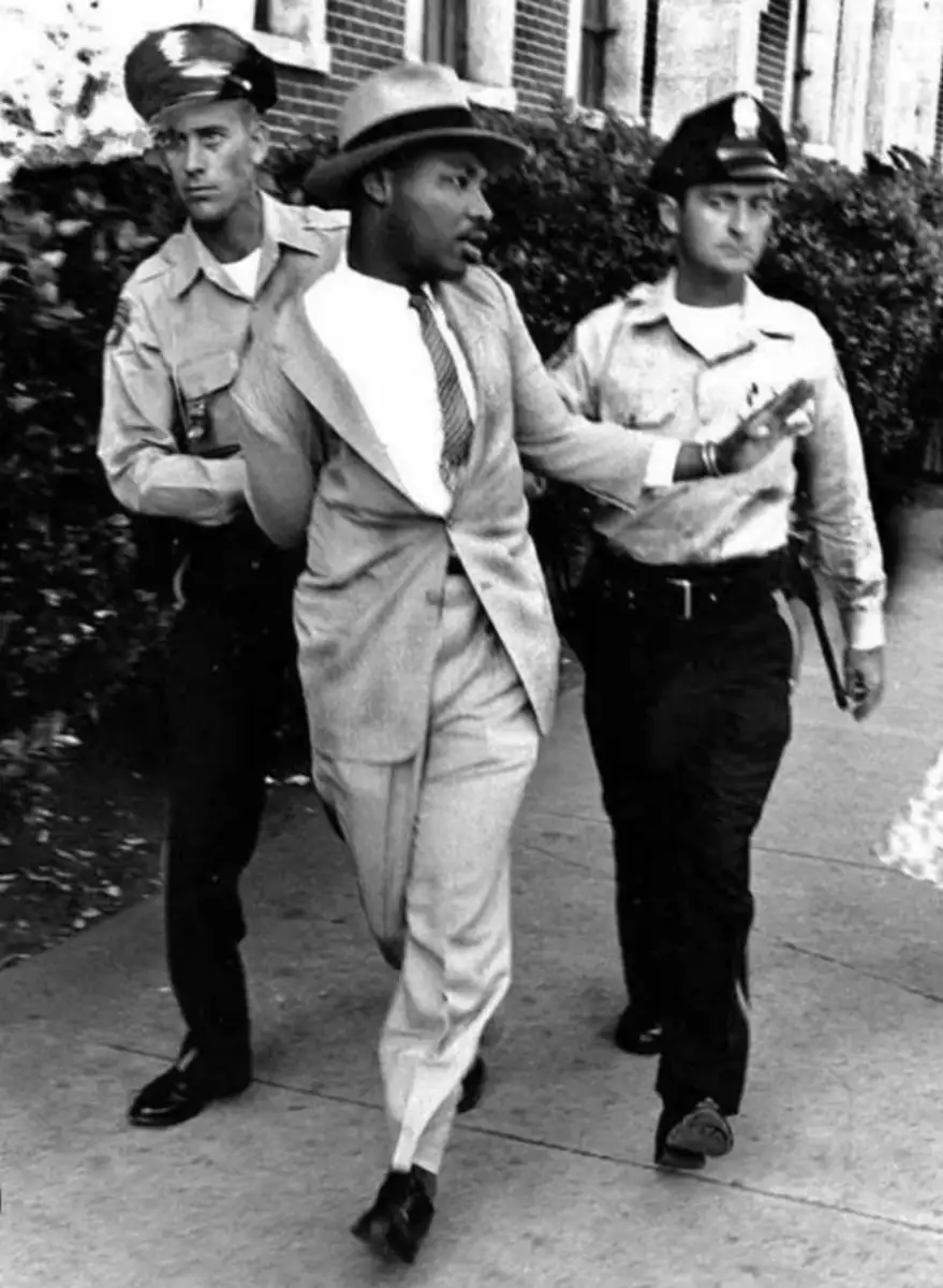 A well-dressed Black man is being escorted by two white police officers on a sidewalk; the man is looking to the side with one arm raised, while the officers hold each of his arms and walk beside him.
