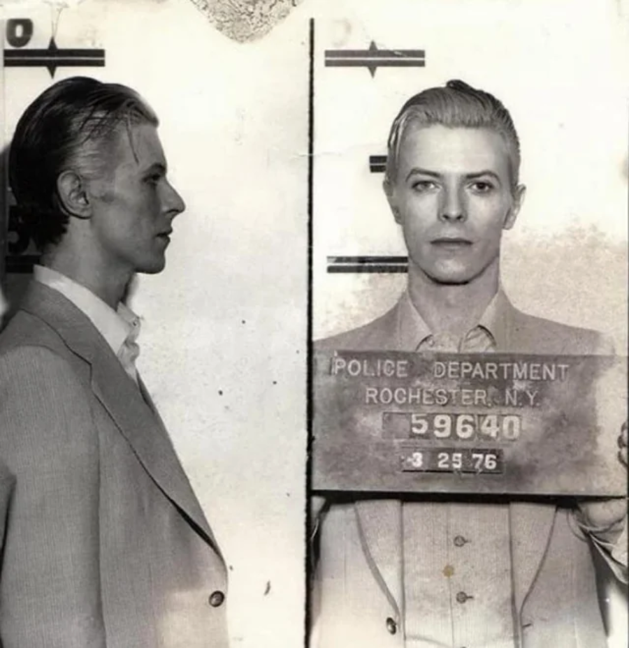 Black and white mugshot photo of a man in a suit with slicked-back hair, shown in both profile and front view, holding a sign that reads "Police Department Rochester, N.Y. 59640 3 25 76.