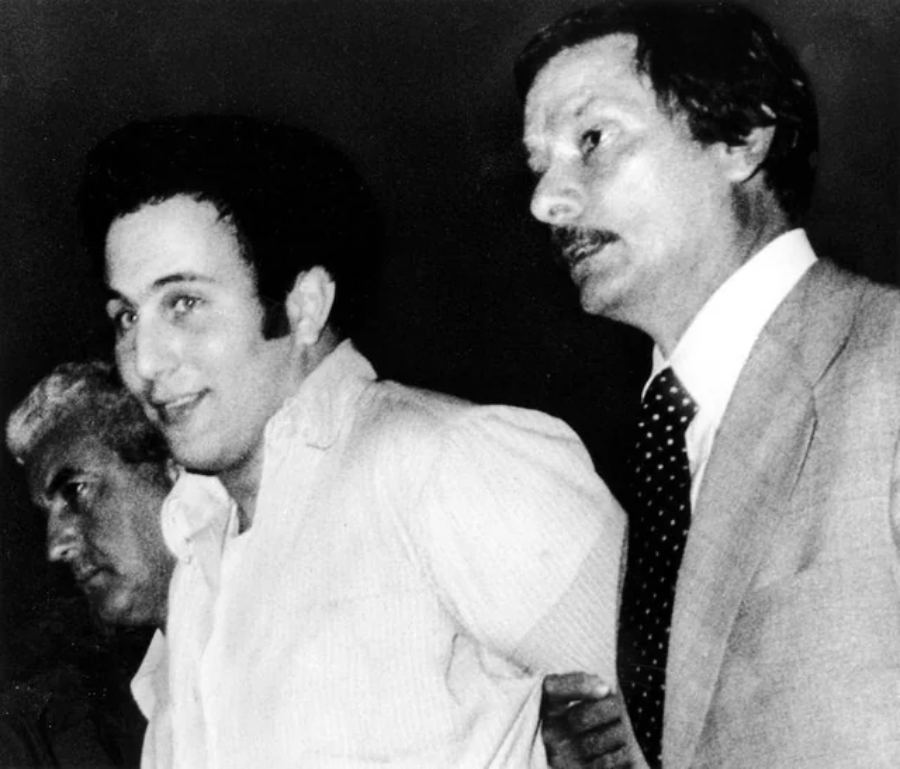 Black and white photo of a man in a light shirt being escorted by two men in suits. The escorted man is looking slightly to the side and smiling, while the others look serious.