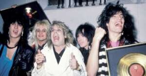 Five men with big, teased 1980s hairstyles celebrate and smile while holding gold records. They wear rock-style outfits, including leather and a white jacket, against a plain indoor background.