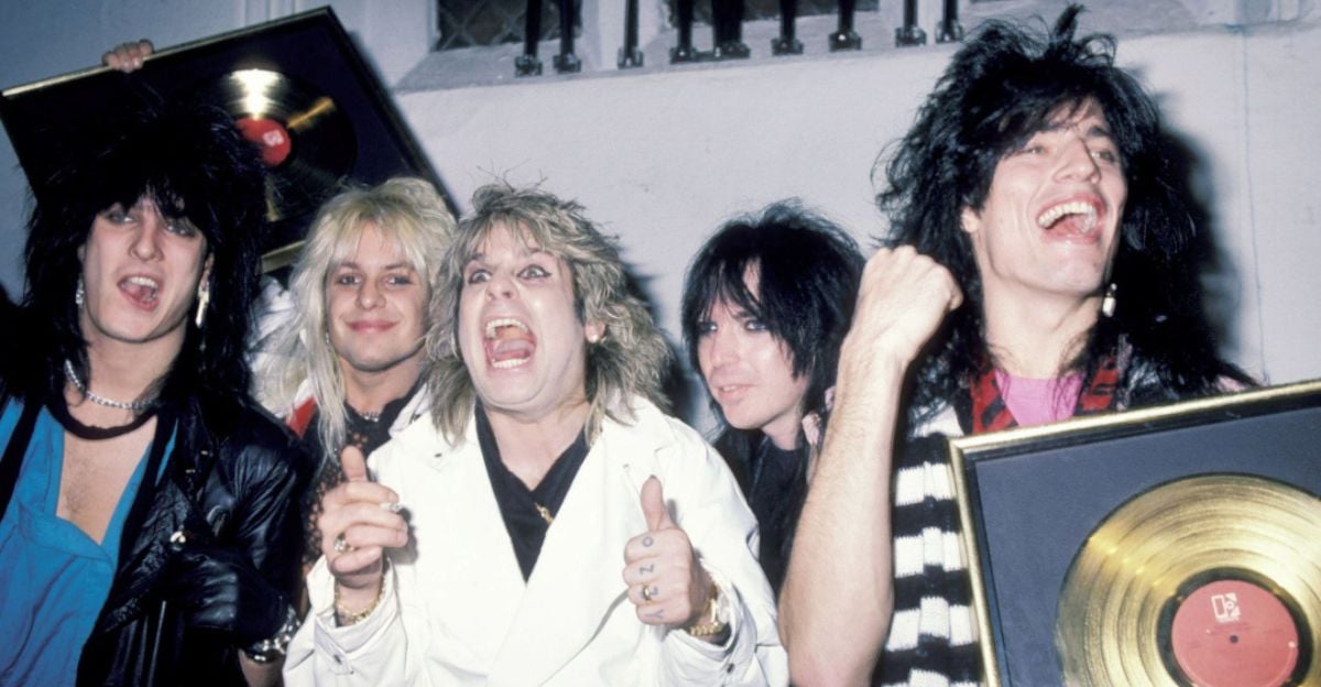 Five men with big, teased 1980s hairstyles celebrate and smile while holding gold records. They wear rock-style outfits, including leather and a white jacket, against a plain indoor background.