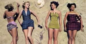 Four women in vintage swimsuits lie on sandy beach. Two hold shoes, one has an arm raised, and a sun hat sits above their heads. The swimsuits are patterned and solid in different colors. They appear relaxed and enjoy the sun.