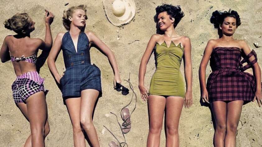 Four women in vintage swimsuits lie on sandy beach. Two hold shoes, one has an arm raised, and a sun hat sits above their heads. The swimsuits are patterned and solid in different colors. They appear relaxed and enjoy the sun.